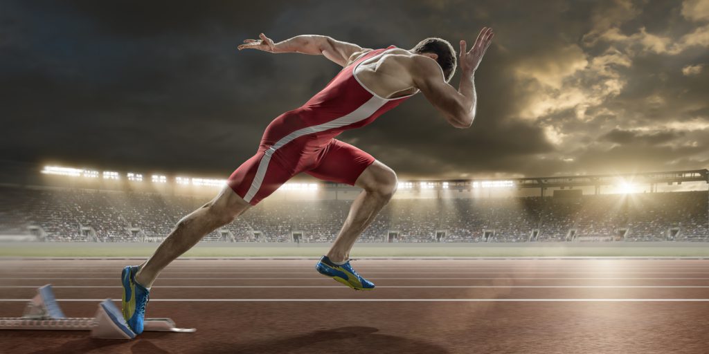 A close up image of a professional powerful male sprinter in mid action sprinting out of starting blocks at high speed, on a running track. The action takes place in a generic outdoor floodlit athletics stadium, full of spectators, under a dramatic evening sky. The athlete wears a bodysuit and running spikes.